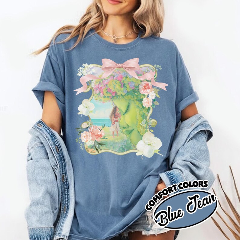 K&ouml;nnte beinhalten: Blaues Blue Jean Comfort Colors T-Shirt mit floralem Design, rosa Schleife und Strandszene. Das T-Shirt hat hochgekrempelte &Auml;rmel und eine lockere Passform. Der Text "COMFORT COLORS Blue Jean" ist unten rechts aufgedruckt.
