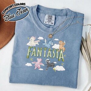 May include: A blue Comfort Colors t-shirt featuring the word "FANTASIA" in green, and a design of cartoon pegasus characters. The shirt has a crew neck and short sleeves. The words "Comfort Colors" and "Blue Jean" are in a circular logo.