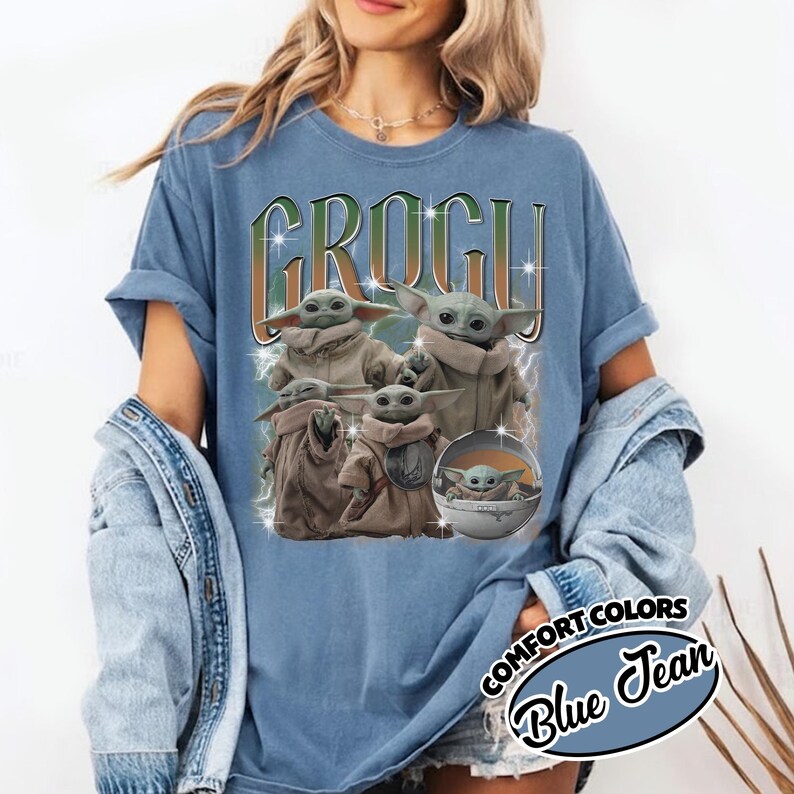 K&ouml;nnte beinhalten: Ein blaues, jeansfarbenes T-Shirt mit einem Grafikdruck des Charakters Grogu in verschiedenen Posen. Der Text GROGU ist in gro&szlig;en, stilisierten Buchstaben &uuml;ber den Figuren abgebildet, erg&auml;nzt durch Sternakzente und Blitzeffekte.
