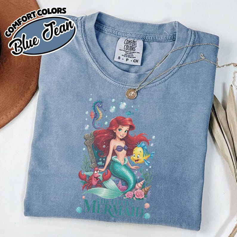 May include: Blue jean Comfort Colors t-shirt featuring a graphic of Ariel from The Little Mermaid, with Flounder and Sebastian. The shirt has the text "The Little Mermaid" at the bottom. A gold necklace is draped across the shirt.