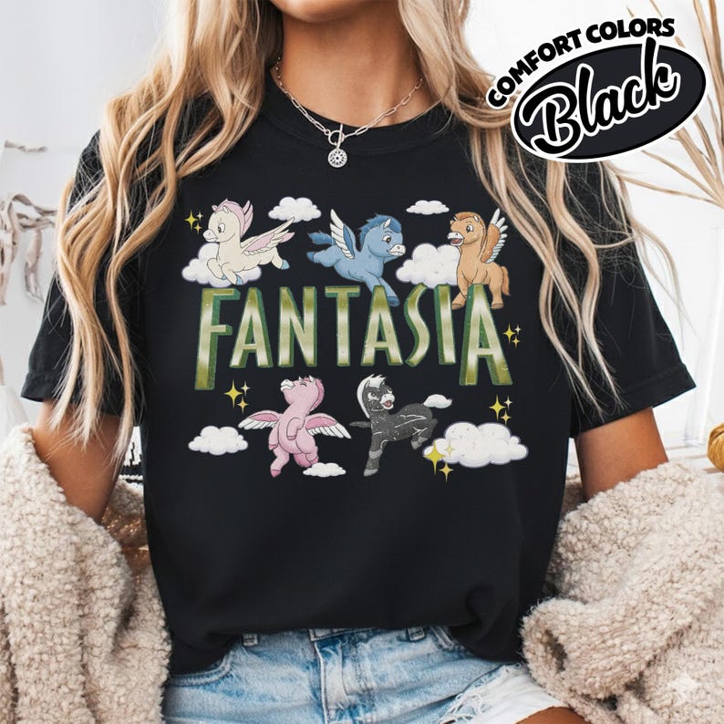 May include: Black t-shirt with the word "FANTASIA" in green and white lettering. The shirt features cartoon pegasus characters in various colours, including pink, blue, and tan, with white clouds and yellow stars. The text "COMFORT COLORS Black" is in a circular design.