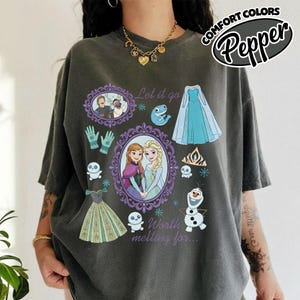 Op de afbeelding: Donkergrijs T-shirt met illustraties uit de film "Frozen". Het ontwerp bevat afbeeldingen van Anna en Elsa, Olaf, en de teksten "Let it go" en "Worth melting for..." in een speels lettertype.