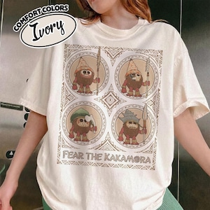 May include: Ivory t-shirt with a graphic of four cartoon characters in circular frames. The characters are brown with red accents, holding spears. The text "FEAR THE KAKAMORA" is below the graphic. The shirt has the text "COMFORT COLORS" and "Ivory" in an oval.