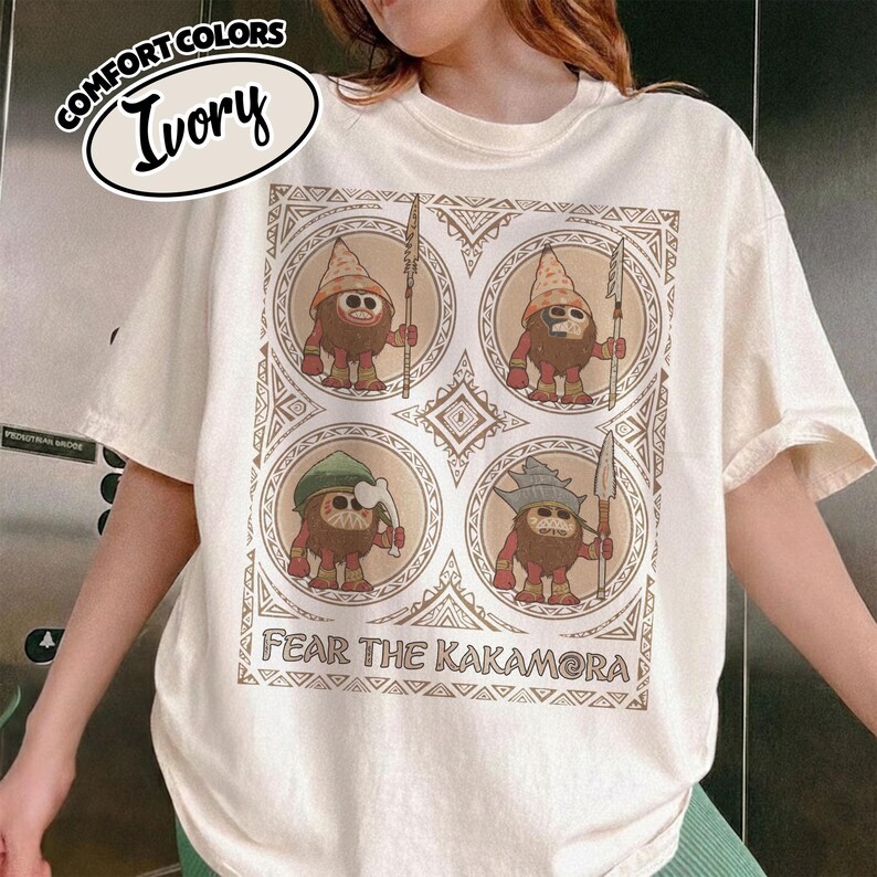 May include: Ivory t-shirt with a graphic of four cartoon characters in circular frames. The characters are brown with red accents, holding spears. The text "FEAR THE KAKAMORA" is below the graphic. The shirt has the text "COMFORT COLORS" and "Ivory" in an oval.