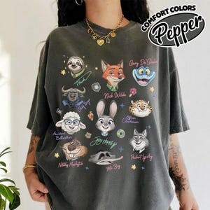 K&ouml;nnte beinhalten: Dunkelgraues T-Shirt mit Cartoon-Charakterportr&auml;ts, darunter Nick Wilde und Judy Hopps. Das Shirt tr&auml;gt die Aufschrift "COMFORT COLORS" und "Pepper". Das T-Shirt hat eine lockere Passform.