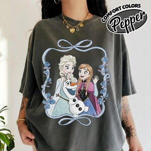 May include: Dark gray t-shirt featuring a graphic of Elsa, Anna, and Olaf from Frozen, framed by a blue ribbon and floral accents. The text "COMFORT COLORS Pepper" is printed on the upper right. The shirt has short sleeves.