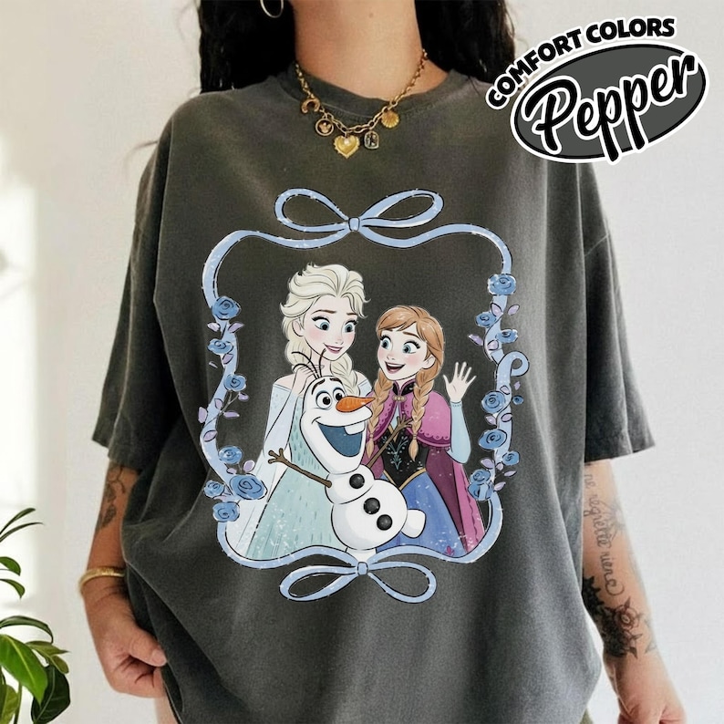 May include: Dark gray t-shirt featuring a graphic of Elsa, Anna, and Olaf from Frozen, framed by a blue ribbon and floral accents. The text "COMFORT COLORS Pepper" is printed on the upper right. The shirt has short sleeves.