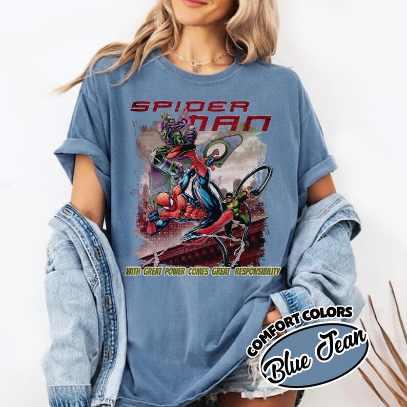 Op de afbeelding: Blauw T-shirt met een Spider-Man-afbeelding. De tekst "SPIDER-MAN" is rood en het shirt heeft een stripboekillustratie van Spider-Man en andere personages. De tekst "WITH GREAT POWER COMES GREAT RESPONSIBILITY" is ook aanwezig.