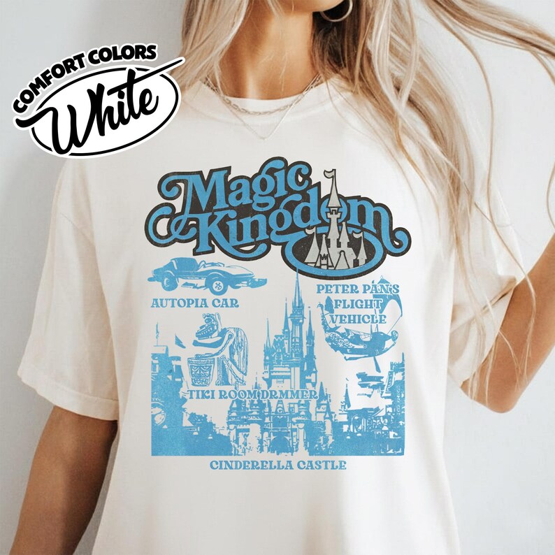May include: White Comfort Colors t-shirt featuring a blue graphic design with the words "Magic Kingdom" and illustrations of attractions like Autopia Car and Cinderella Castle. The shirt has short sleeves and a crew neck.