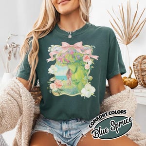 K&ouml;nnte beinhalten: Blau-Fichte Comfort Colors T-Shirt mit floralem Design, rosa Schleife und Landschaftsillustration. Das Design umfasst Blumen, eine Person und eine Strandszene. Das Shirt hat kurze &Auml;rmel.