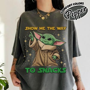 May include: Dark gray t-shirt featuring a cartoon image of Grogu, also known as Baby Yoda, pointing upwards. The text "SHOW ME THE WAY TO SNACKS" is printed in yellow and green. The shirt has a relaxed fit and short sleeves.