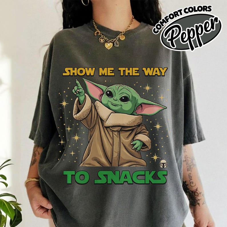 May include: Dark gray t-shirt featuring a cartoon image of Grogu, also known as Baby Yoda, pointing upwards. The text "SHOW ME THE WAY TO SNACKS" is printed in yellow and green. The shirt has a relaxed fit and short sleeves.