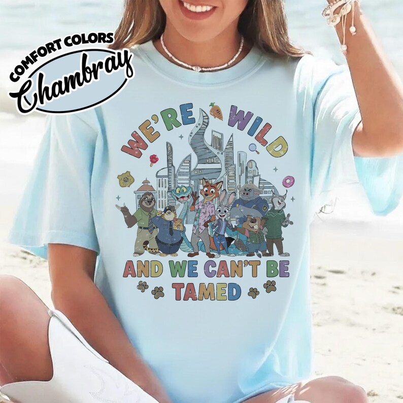 May include: Light blue Comfort Colors Chambray t-shirt with the text "WE'RE WILD AND WE CAN'T BE TAMED" and a colorful graphic of cartoon animals in front of a city skyline. The shirt has short sleeves.