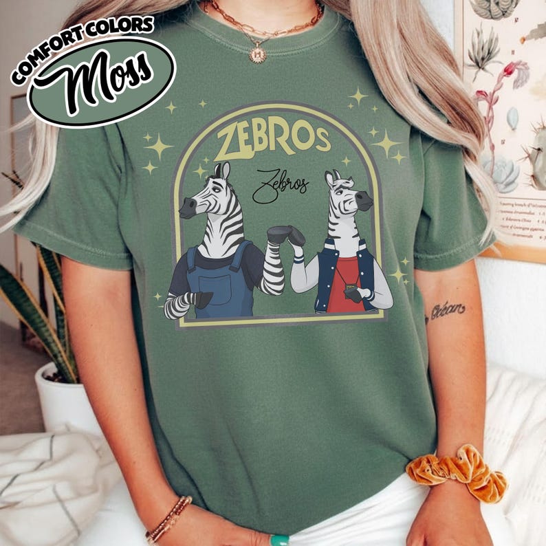 May include: A moss green t-shirt featuring a graphic of two zebras within a yellow arch, with the text "ZEBROS" above and "Zebros" below. One zebra is in overalls, the other in a jacket. The shirt also has the text "COMFORT COLORS" and "Moss" in a circle.