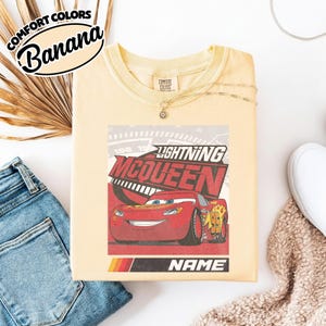 May include: A pale yellow t-shirt featuring a graphic of the red race car Lightning McQueen with the text Lightning McQueen and Name. The shirt has a crew neckline and a Comfort Colors brand label at the inner neck. The design includes a speedometer background.