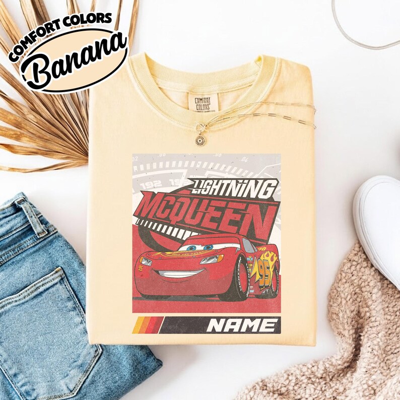 May include: A pale yellow t-shirt featuring a graphic of the red race car Lightning McQueen with the text Lightning McQueen and Name. The shirt has a crew neckline and a Comfort Colors brand label at the inner neck. The design includes a speedometer background.