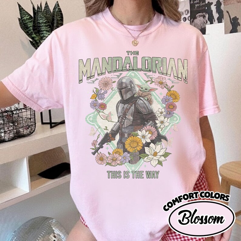 May include: A light pink t-shirt featuring a graphic of The Mandalorian and Grogu surrounded by colorful flowers. The text reads THE MANDALORIAN at the top and THIS IS THE WAY at the bottom. The design is set against a light green diamond shape.