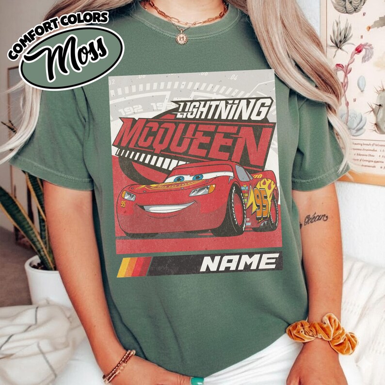 May include: A moss green t-shirt featuring a graphic of the red race car Lightning McQueen with the text Lightning McQueen and Name. The design includes a speedometer background, yellow and orange flames, and the number 95 on the car side.
