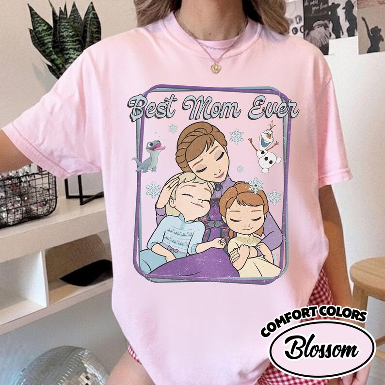 May include: Pink Comfort Colors t-shirt with a graphic featuring the phrase "Best Mom Ever" and a cartoon image of a mother and two children. The design includes snowflakes and a cartoon snowman. The shirt is a light pink color.