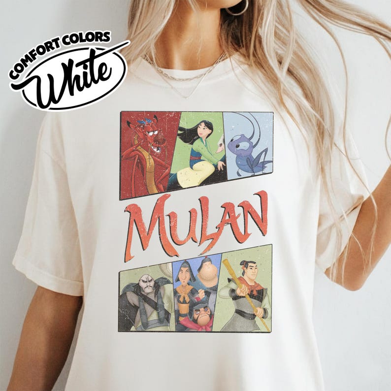 Puede incluir: Camiseta blanca Comfort Colors con un gr&aacute;fico de personajes de Mulan al estilo c&oacute;mic. La palabra "Mulan" est&aacute; en letras naranjas. La camiseta tiene un aspecto vintage y desgastado.