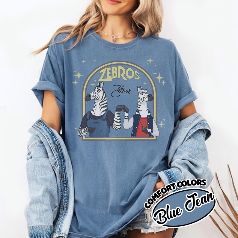 May include: Blue Comfort Colors t-shirt featuring a graphic of two zebras in a gold arch with the text "ZEBROS" and "Zebros". One zebra wears overalls, the other a jacket. The shirt is a blue jean colour.