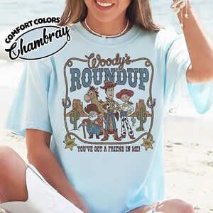 May include: Light blue t-shirt with a graphic featuring characters from "Woody's Roundup." The design includes Woody, Jessie, and other characters, with the text "Woody's ROUNDUP" and "YOU'VE GOT A FRIEND IN ME!" printed on the front. The shirt has the "Comfort Colors" and "Chambray" logos.