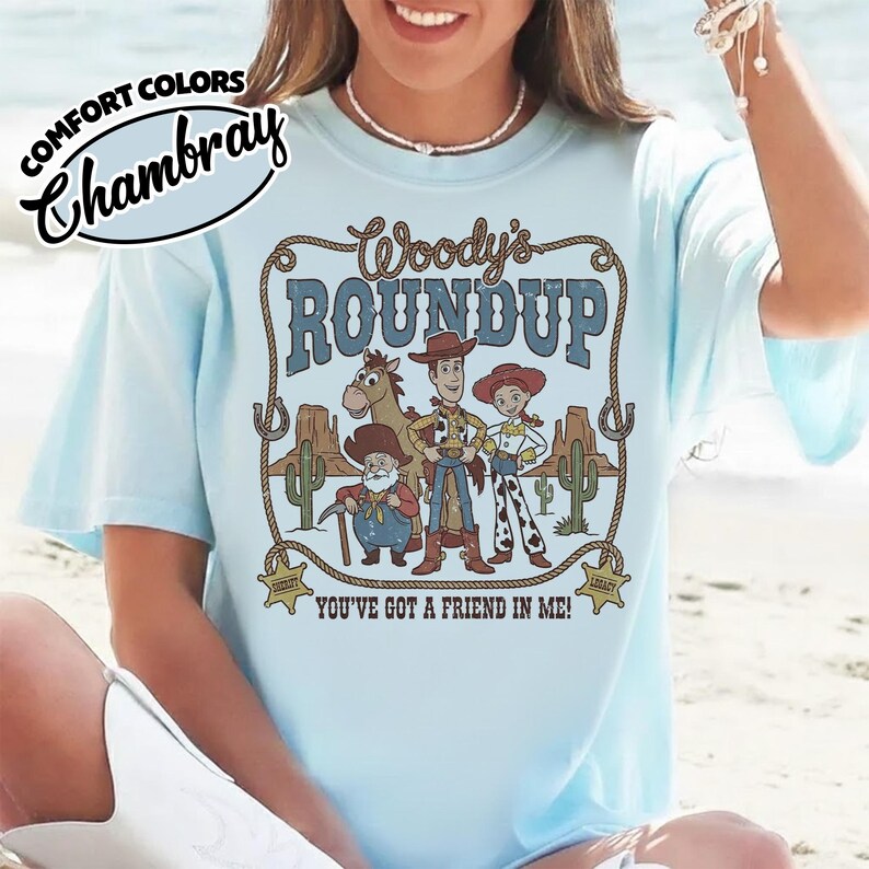 May include: Light blue t-shirt with a graphic featuring characters from "Woody's Roundup." The design includes Woody, Jessie, and other characters, with the text "Woody's ROUNDUP" and "YOU'VE GOT A FRIEND IN ME!" printed on the front. The shirt has the "Comfort Colors" and "Chambray" logos.