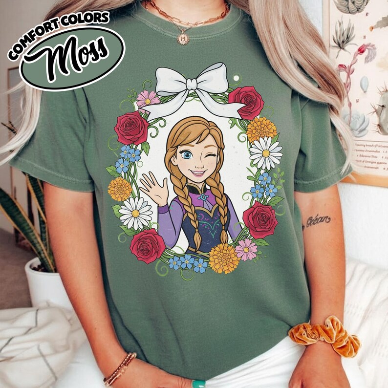 Peut inclure: T-shirt vert mousse avec une image de dessin anim&eacute; d'un personnage aux tresses blondes, faisant un clin d'&oelig;il et saluant. Le personnage est encadr&eacute; par une couronne florale avec des roses rouges, des marguerites et des fleurs bleues. Les mots "COMFORT COLORS" et "Moss" sont dans un design circulaire.