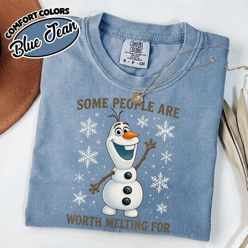 May include: Blue jean Comfort Colors t-shirt featuring a graphic of Olaf the snowman with the text "SOME PEOPLE ARE WORTH MELTING FOR." The shirt has a crew neck and short sleeves. A silver necklace is draped across the shirt.
