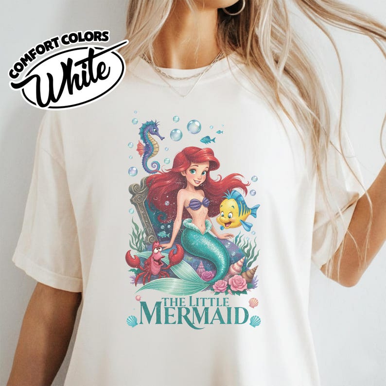 May include: White Comfort Colors t-shirt featuring a colorful graphic of Ariel from The Little Mermaid. The design includes Ariel, Flounder, Sebastian, and a seahorse, with the words "The Little Mermaid".