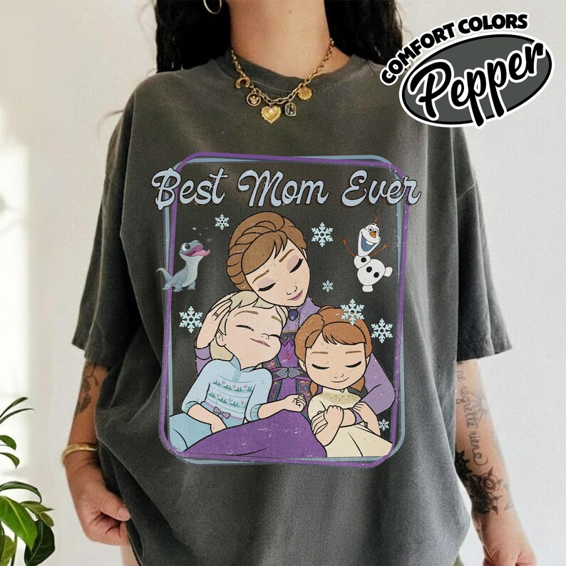 May include: Dark gray t-shirt with a graphic that says "Best Mom Ever." The graphic features a mother and two children, with snowflakes and cartoon characters. The shirt has the words "Comfort Colors Pepper" in a circle.