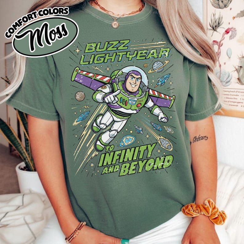 May include: A moss green t-shirt featuring a graphic of Buzz Lightyear in a space suit with purple wings. The text reads Buzz Lightyear to infinity and beyond. The design includes small planets, stars, and rockets in a cartoon style on the front.