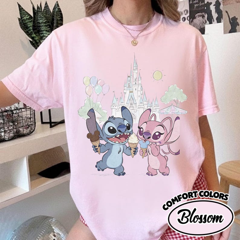 Puede incluir: Camiseta rosa claro con un dibujo animado de Stitch y Angel sosteniendo helados frente a un castillo. La camiseta tiene las palabras "COMFORT COLORS" y "Blossom" impresas.