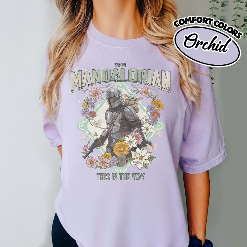 May include: A light purple t-shirt featuring a graphic print of The Mandalorian and Grogu surrounded by colorful flowers and butterflies. The text reads THE MANDALORIAN at the top and THIS IS THE WAY at the bottom. A Comfort Colors Orchid label is visible.