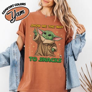 May include: A rust-colored t-shirt with the text "SHOW ME THE WAY TO SNACKS" and a graphic of a green alien character. The shirt also features the words "COMFORT COLORS" and "Yam" in a logo.