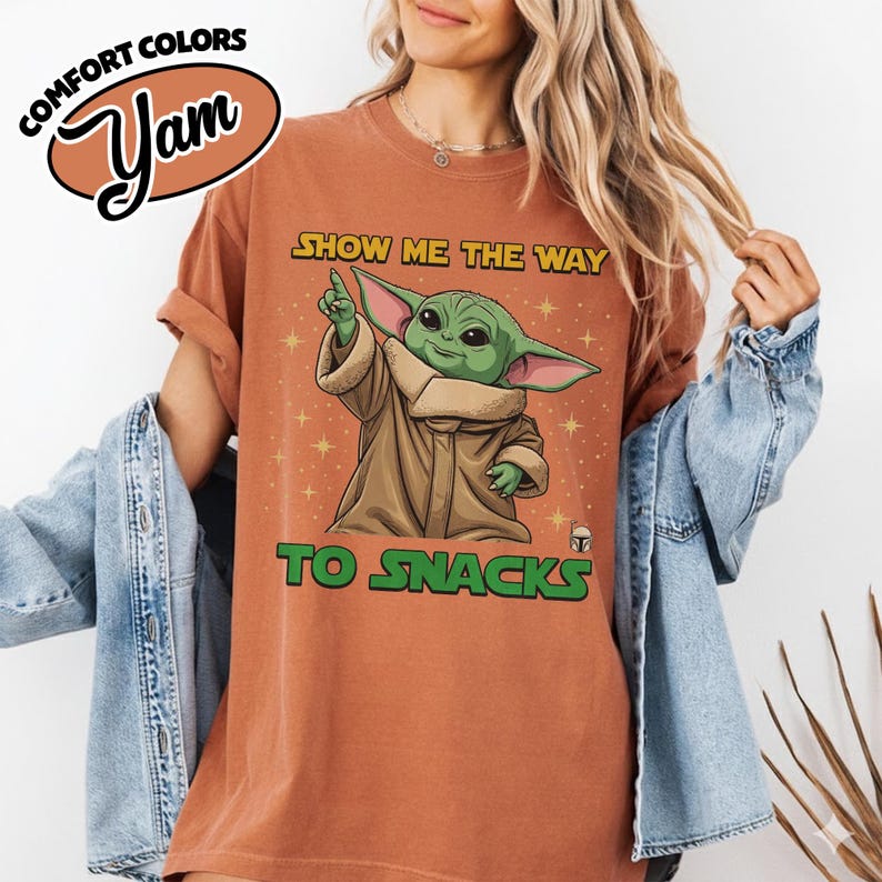 May include: A rust-colored t-shirt with the text "SHOW ME THE WAY TO SNACKS" and a graphic of a green alien character. The shirt also features the words "COMFORT COLORS" and "Yam" in a logo.