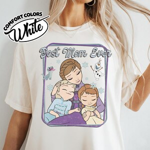 May include: White Comfort Colors t-shirt featuring a graphic of a mother and two children with the text "Best Mom Ever". The graphic includes cartoon characters and snowflakes. The shirt has short sleeves.