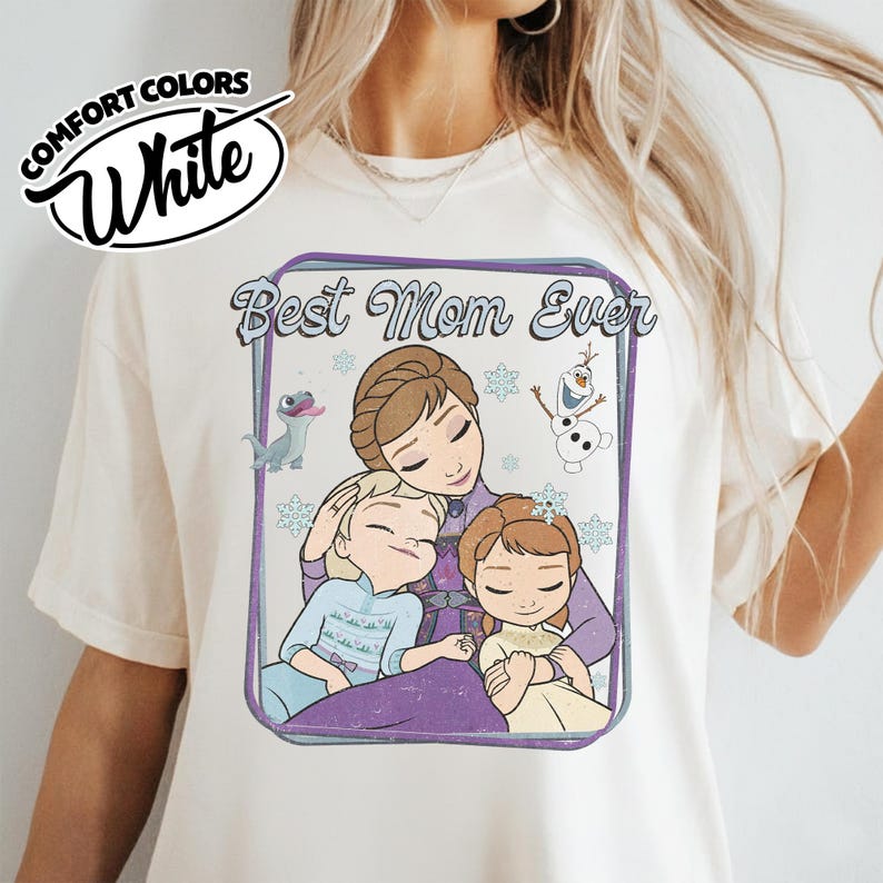 May include: White Comfort Colors t-shirt featuring a graphic of a mother and two children with the text "Best Mom Ever". The graphic includes cartoon characters and snowflakes. The shirt has short sleeves.