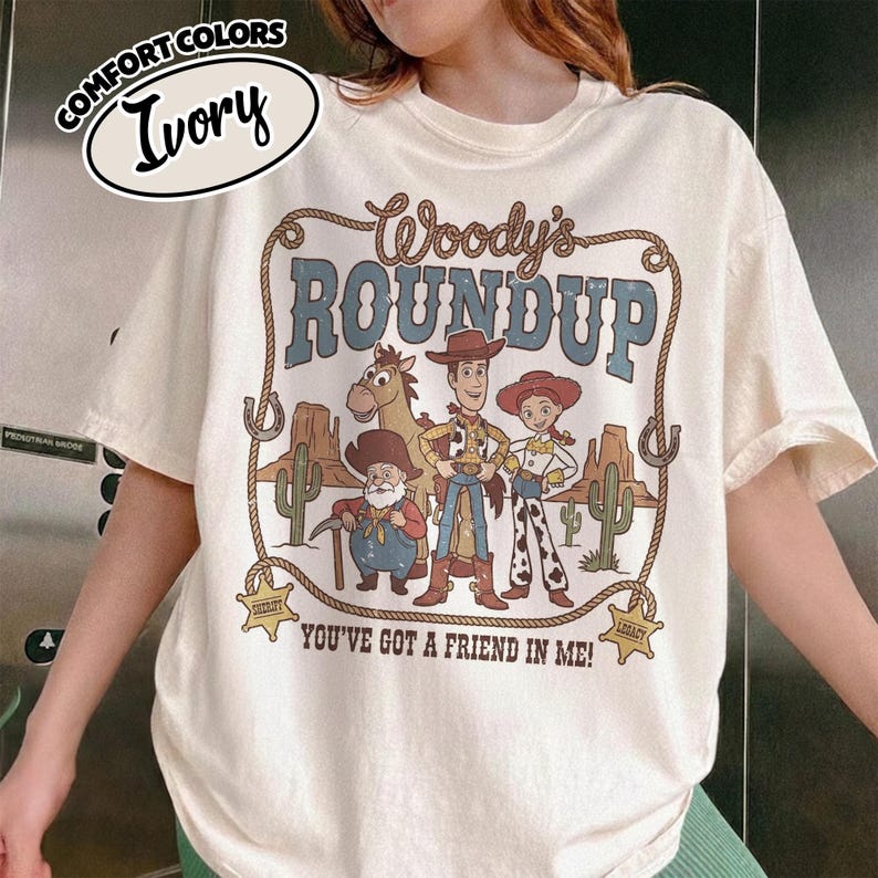 May include: Ivory-colored t-shirt featuring a graphic of Woody, Jessie, and other characters from Woody's Roundup. The design includes the text "Woody's Roundup" and "You've Got a Friend in Me!" in a Western-style font. The shirt has a crew neck and short sleeves.