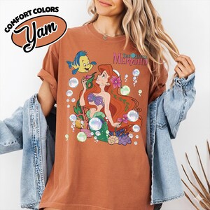 K&ouml;nnte beinhalten: Ein rostfarbenes Kurzarm-T-Shirt mit einem Grafikdruck von Arielle und Fabius aus Arielle, die Meerjungfrau, umgeben von Blasen, Korallen und Muscheln. Der Text Comfort Colors Yam erscheint in der oberen linken Ecke des Bildes.