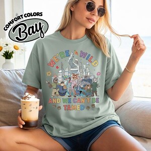 May include: Sage green t-shirt with the text "WE'RE WILD AND WE CAN'T BE TAMED" and a colorful graphic of cartoon characters in front of a city skyline. The shirt also has the Comfort Colors Bay logo.