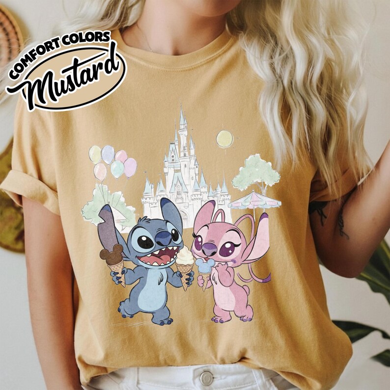 Puede incluir: Camiseta de color mostaza con una ilustraci&oacute;n de dibujos animados de Stitch y Angel sosteniendo conos de helado frente a un castillo. La camiseta tiene las palabras "COMFORT COLORS" y "Mustard" impresas. Globos y &aacute;rboles pastel tambi&eacute;n est&aacute;n en el fondo.