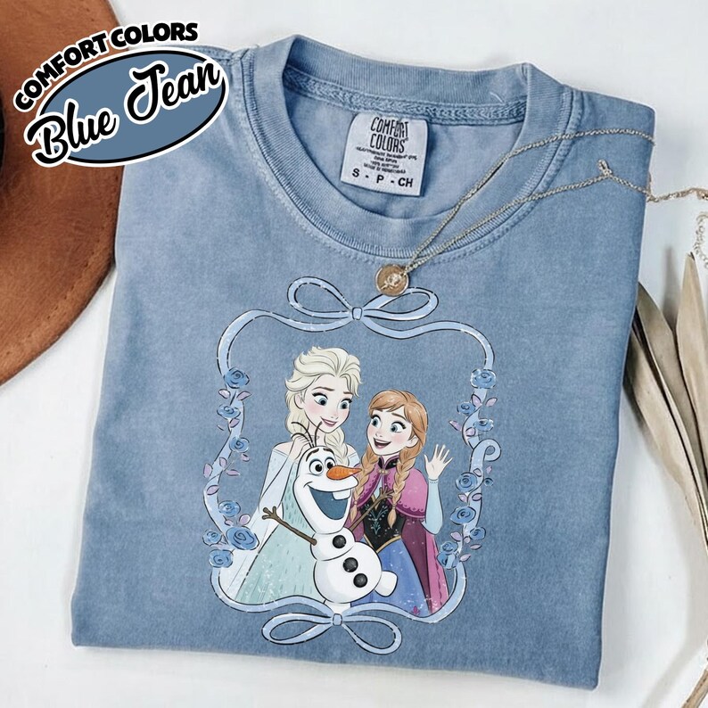 May include: Blue jean colored t-shirt with a graphic of Elsa, Anna, and Olaf from Frozen. The graphic is framed with a blue floral design. The shirt has the words "Comfort Colors" and "Blue Jean" printed on it.