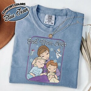 May include: A blue Comfort Colors t-shirt with the words "Best Mom Ever" and a graphic of a mother and two children. The shirt has a crew neck and short sleeves. A silver necklace with a pendant is draped on the shirt.