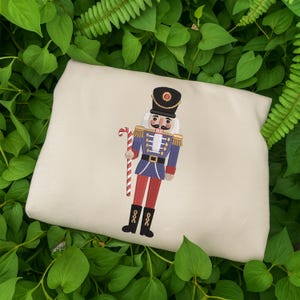 Peut inclure: Un coussin crème avec une broderie d'un soldat casse-noisette tenant une canne en bonbon. Le casse-noisette porte une veste bleue, un pantalon rouge et des bottes noires. Le coussin est entouré de feuilles vertes.