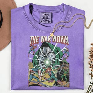 May include: A purple Comfort Colors t-shirt featuring a comic book graphic with the text "THE WAR WITHIN." The graphic depicts various superheroes in action. A gold star pendant necklace rests on the shirt.