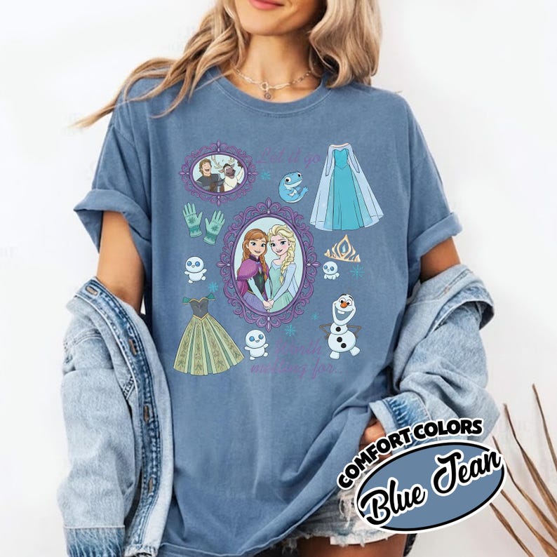 Op de afbeelding: Blauw spijkerbroek Comfort Colors T-shirt met illustraties van personages en elementen uit een populaire animatiefilm. Het ontwerp bevat afbeeldingen van twee zussen, een sneeuwpop en een jurk. De tekst "Let it go" en "making for" is ook zichtbaar.