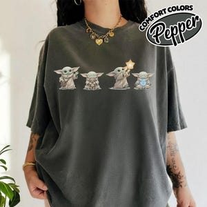 以下が含まれることがあります： 人気のSFシリーズのキャラクターのグラフィックが特徴の濃い灰色のTシャツ。キャラクターは4つの異なるポーズで描かれ、さまざまなアイテムを持っています。「COMFORT COLORS Pepper」の文字がTシャツの右上にプリントされています。