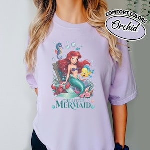 May include: Orchid-colored t-shirt featuring a graphic of Ariel from The Little Mermaid. The design includes Ariel, a seahorse, a crab, and a yellow fish. The words "THE LITTLE MERMAID" are printed at the bottom of the graphic.