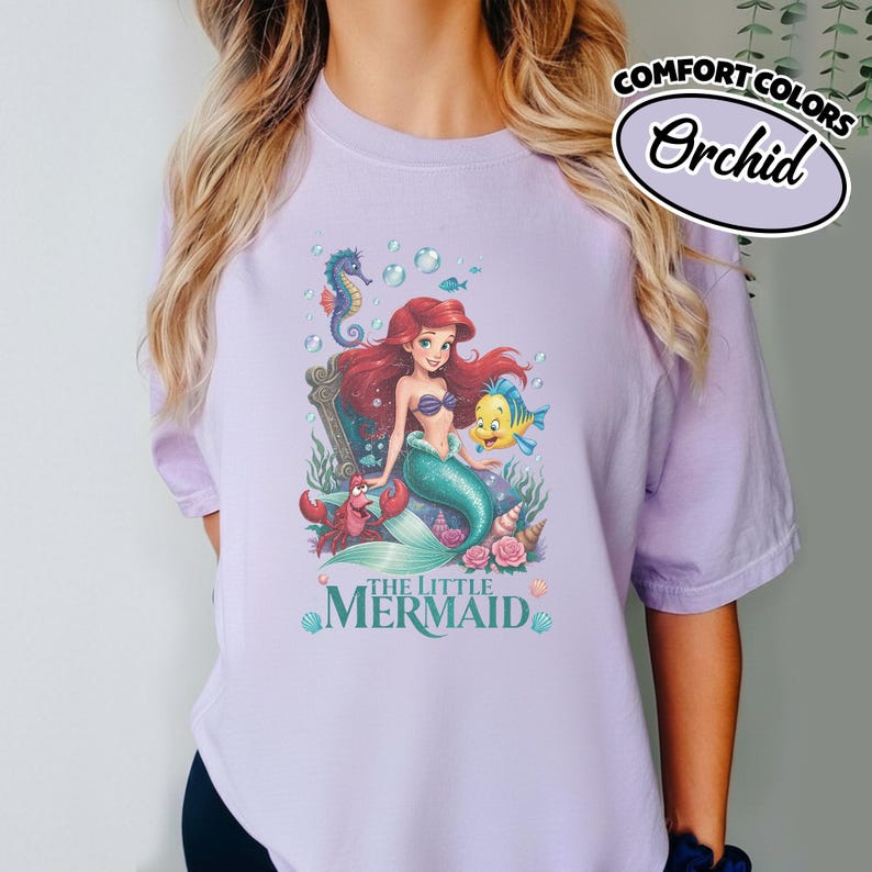 May include: Orchid-colored t-shirt featuring a graphic of Ariel from The Little Mermaid. The design includes Ariel, a seahorse, a crab, and a yellow fish. The words "THE LITTLE MERMAID" are printed at the bottom of the graphic.